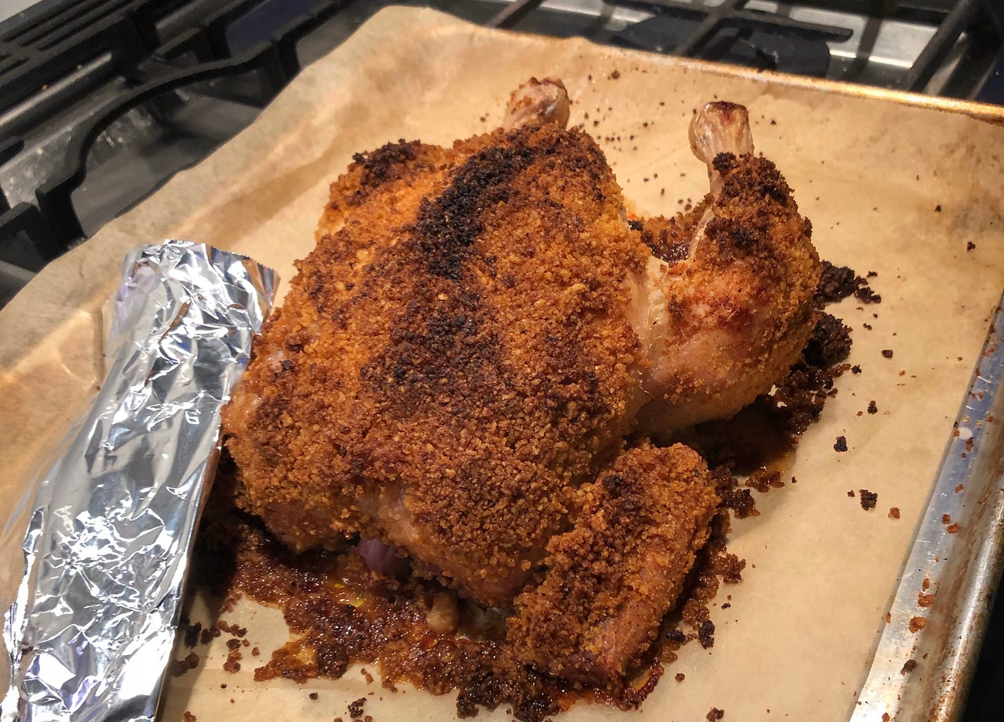 A fully cooked whole chicken, covered in browned potato chip crumbs. It is sitting on a parchment covered sheet pan. 