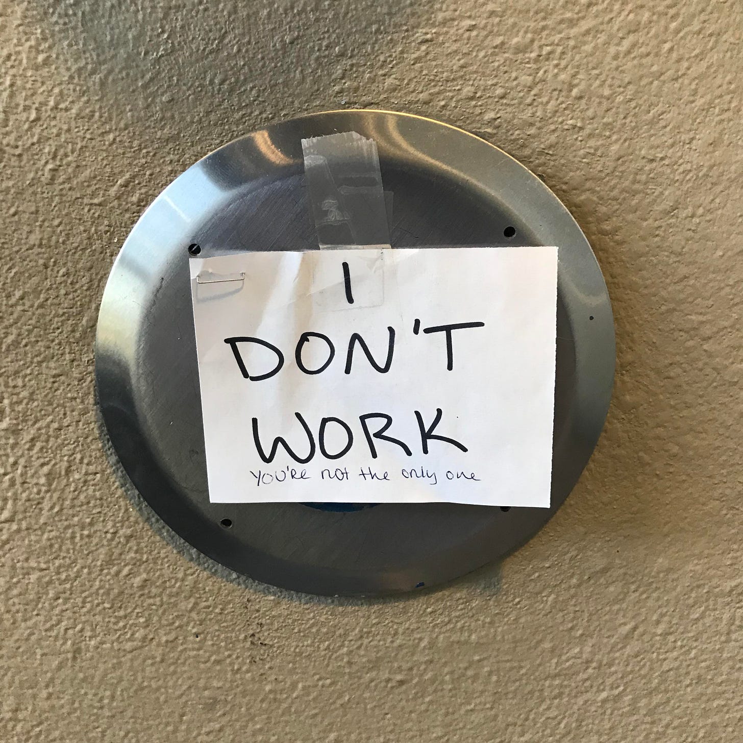 A door button with a piece of paper taped over it that says "I DON'T WORK" in marker. Written underneath it in different, smaller handwriting are the words "You're not the only one."