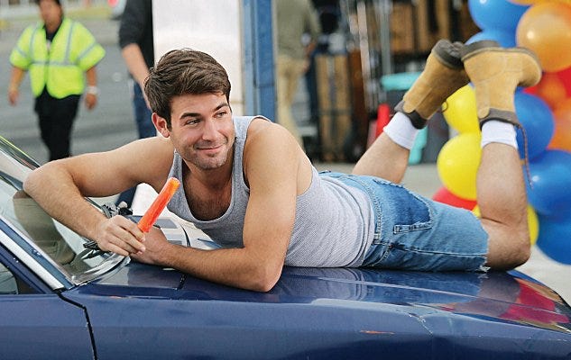 James Wolk in a scene from the television show The Crazy Ones. He is posing atop a car holding an iceblock while wearing jean cut-off shorts and looking off camera with a sexy expression.