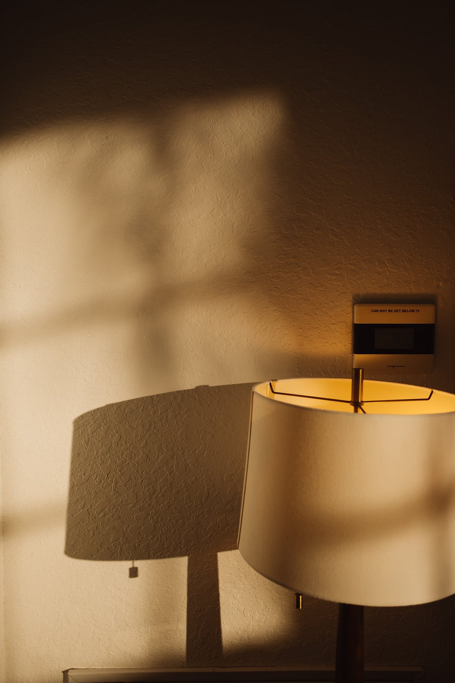 Sunset indoors; the golden light casting dappled shadows on the wall. A lamp switched on and the thermostat that says "CAN NOT BE SET BELOW 72"