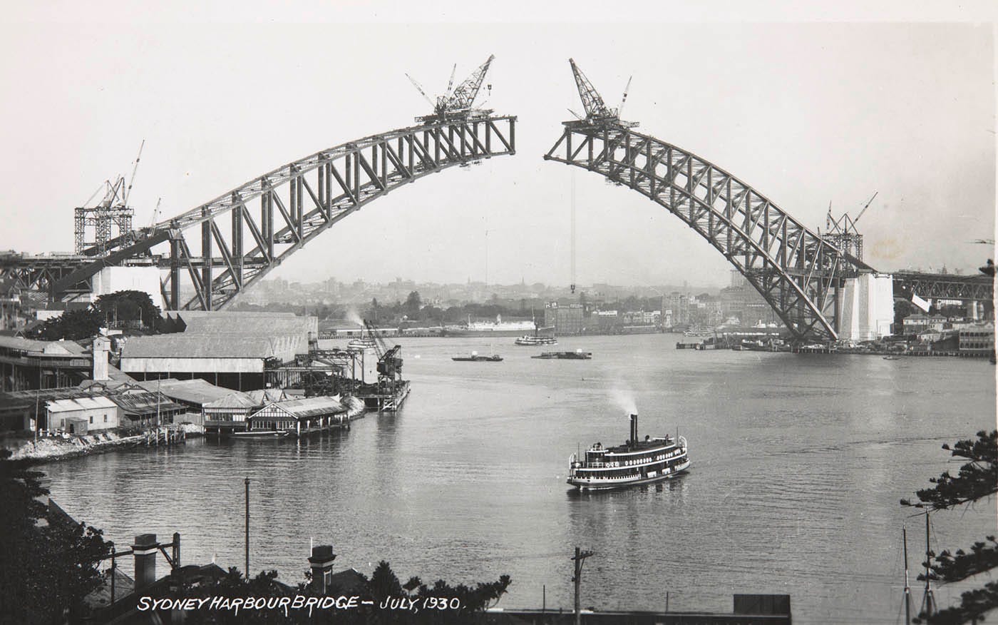 Photo taken from the north shore showing the two sides of the arch under construction. They have yet to meet. Large cranes are perched on top of each end of the spans. A ferry can be seen in the foreground.