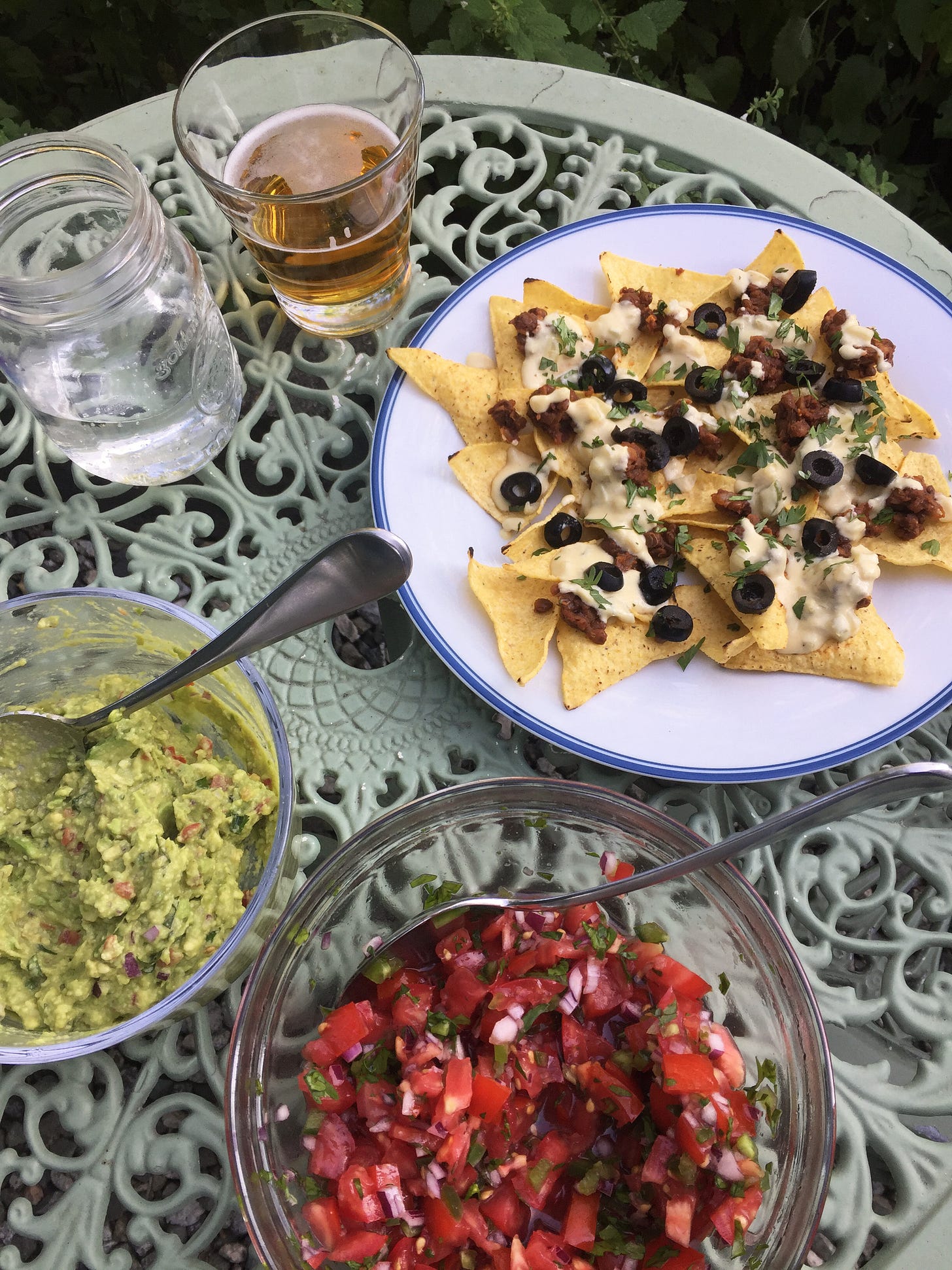 a wrought-iron table with a plate of nachos next to bowls of guacamole and pico de gallo. On the nachos is a cheezy sauce, sliced black olives, and cilantro. A spoon sits in each bowl.