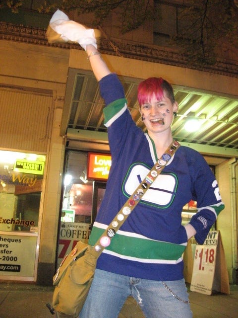 a photo of a younger me sporting a pink mohawk in a Canucks jersey, waving a white towel, grinning with a cigar between my teeth. It is evening and I am standing on the sidewalk in front of the red backlit sign for Love at First Bite pizza.