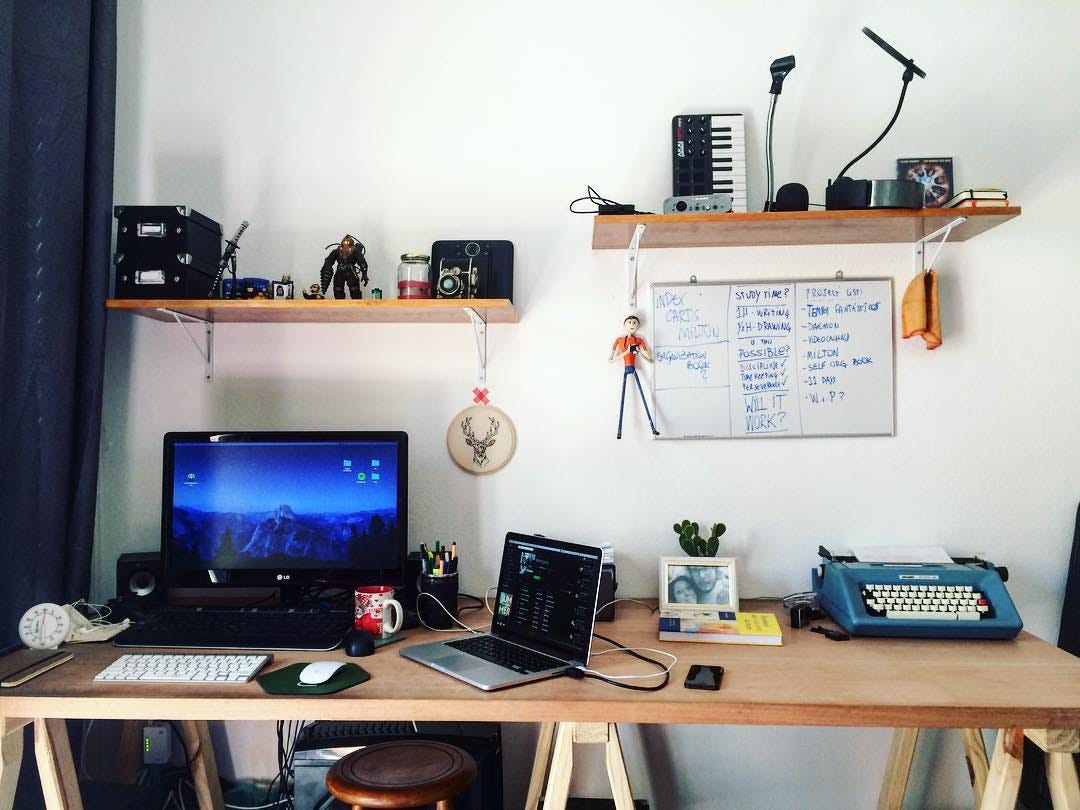 A picture of a work table with an open laptop with an external monitor, as well as a writing machine. There are other things on this table: notebooks, keyboard, mouse, small cactus, picture frame, and others.