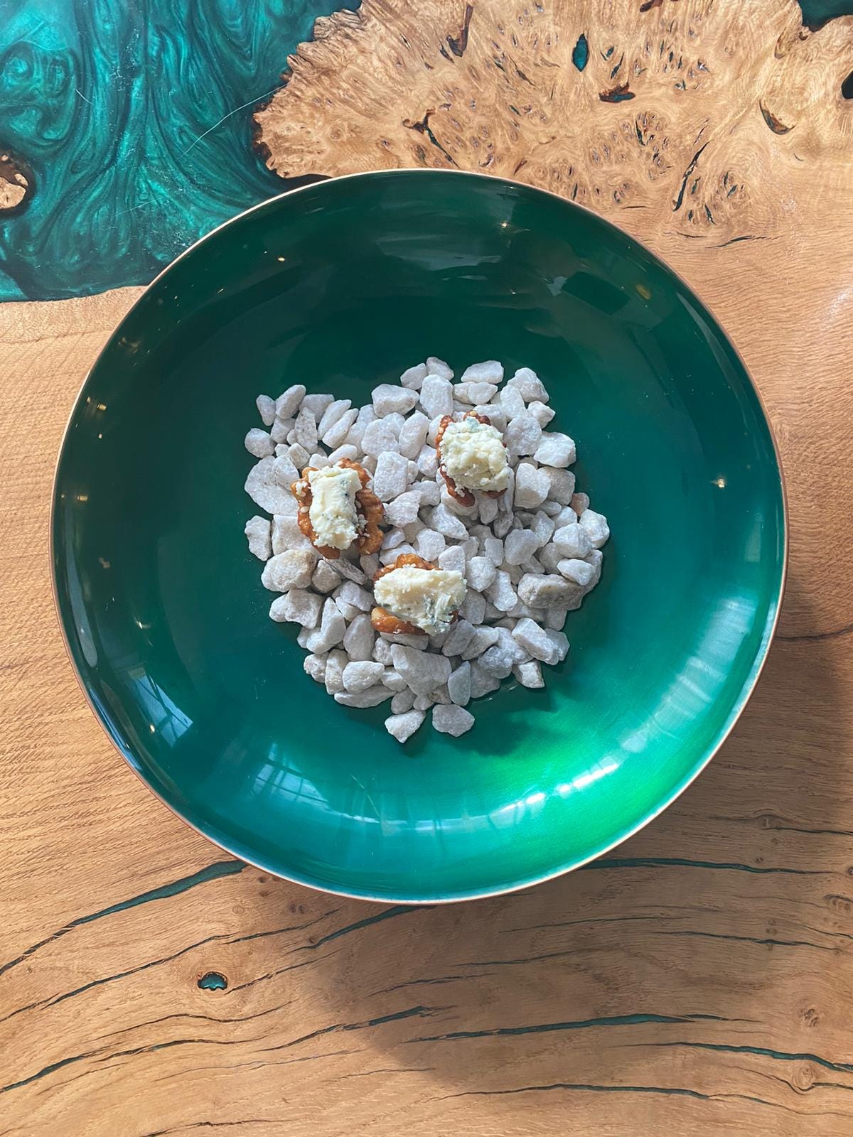 an overhead image of a round turquoise dish in the middle of which is a pile of a small grey stones - on top of the stones sits three walnuts, evenly spaced, with a dollop of cheese on top.