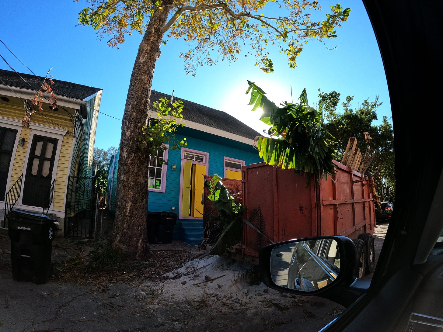 the sun is shining on a colorful blue house with yellow windows as bananna tree stretches out over a dumpster - this is classic shot of Bywater neighborhood in new orleans