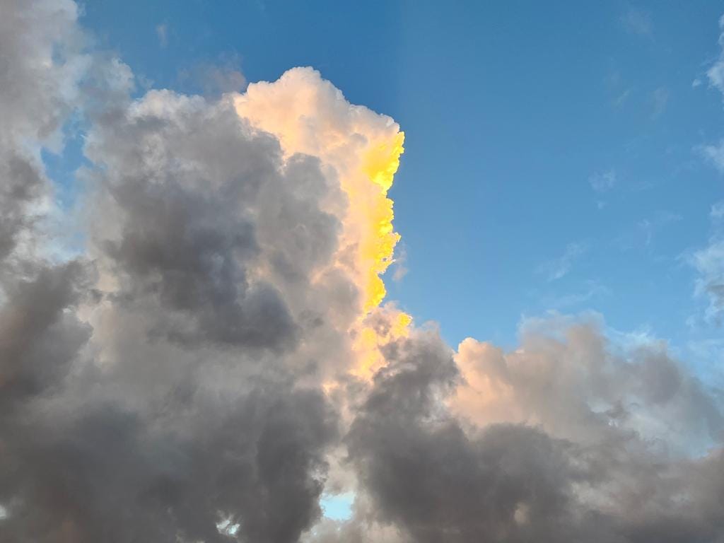 Clouds float along a bright blue sky, with a bright flash of yellow from the sunset illuminating the center of the clouds.