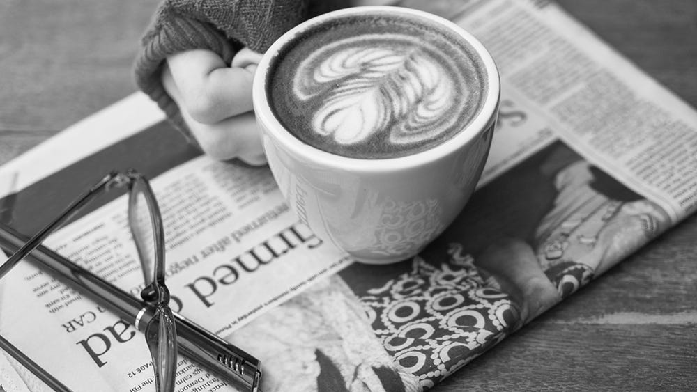 A hand holds a coffee cup with latte art, resting on a folded newspaper. Via Pixabay.