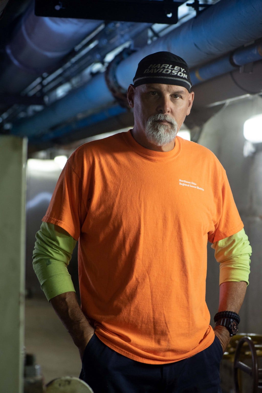 Man with a goatee, Harley Davidson beanie cap, and bright orange NEORSD maintenance shirt stands in a dimly lit hallway deep in the bowels of the Easterly treatment plant. He has hands in the pockets of his dark blue work work pants and making stoic eye contact with the camera.