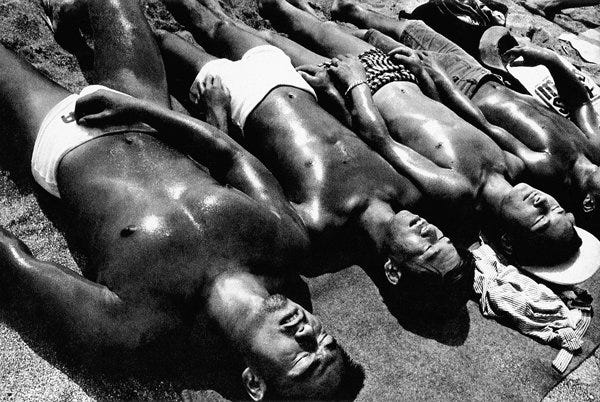 Fotografia preto e branco de vários homens japoneses deitados enfileirados, tomando um bronze de sunga. Eles estão de olhos fechados e o contraste da luz sugere que o corpo deles está úmido. Os corpos foram fotografados com a cabeça virada para baixo, e dispostos em um ângulo apontando para a diagonal direita