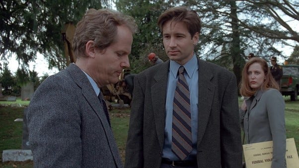 A grey haired man looking downwards, with Mulder looking at him and Scully, much shorter than both, in the background.