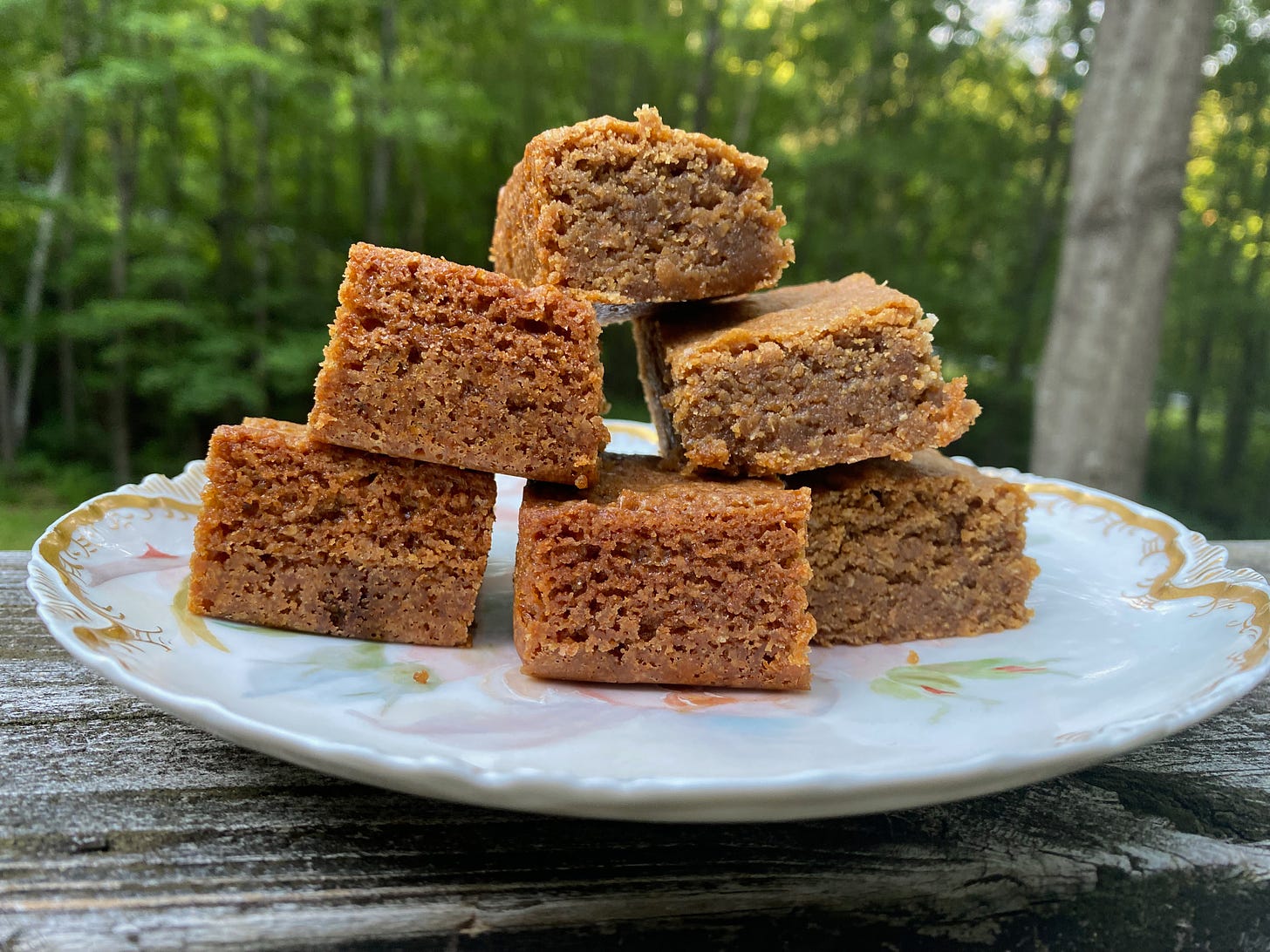 Seven butterscotch brownies stacked in a small pyramid on a ceramic plate on porch railing. Blurry trees are visible in the background.