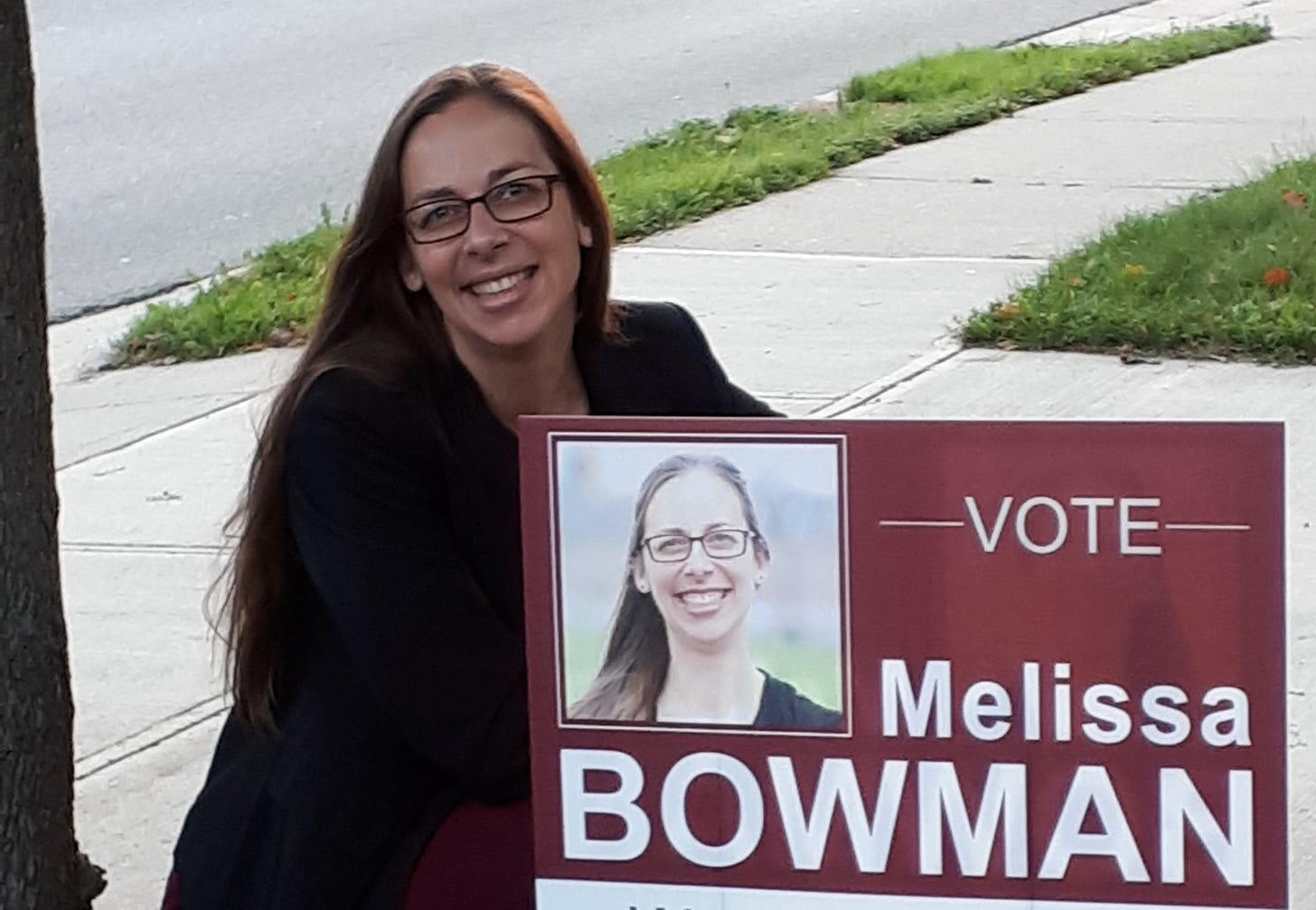 Woman (Melissa Bowman) kneeling down beside an "Vote Melissa Bowman' election sign.