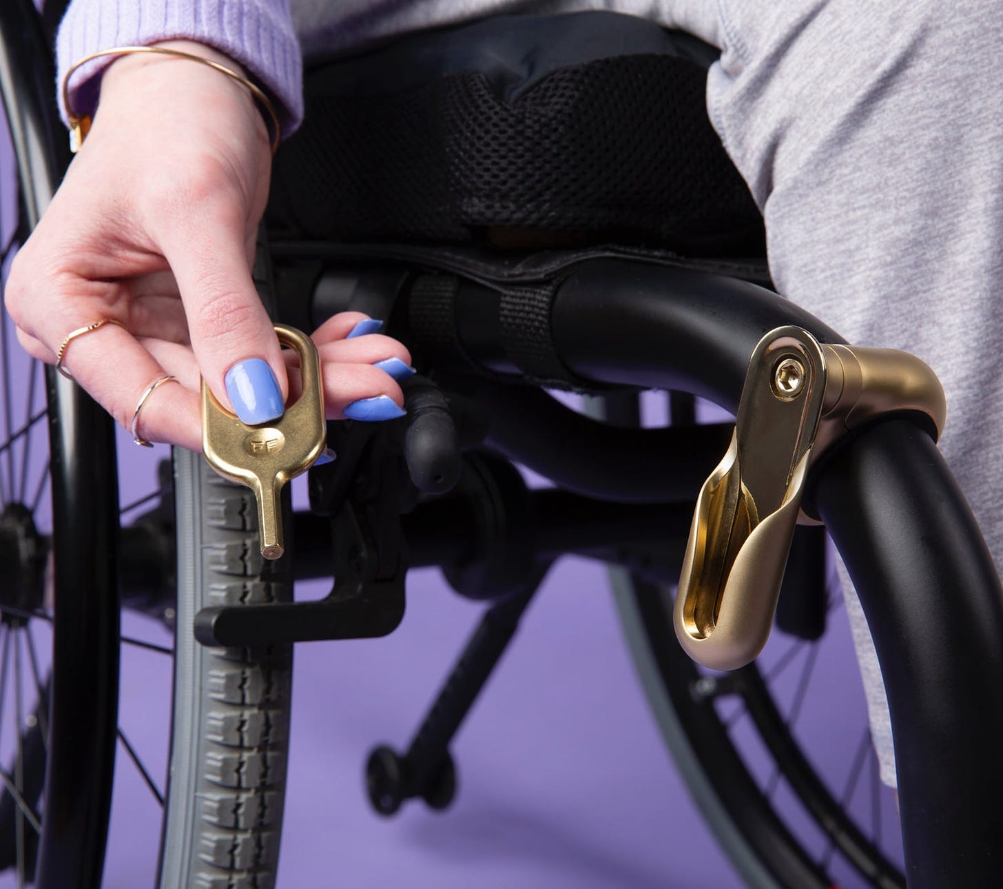 A femme hand with painted nails holds a golden key next to an attachment base on a manual wheelchair.