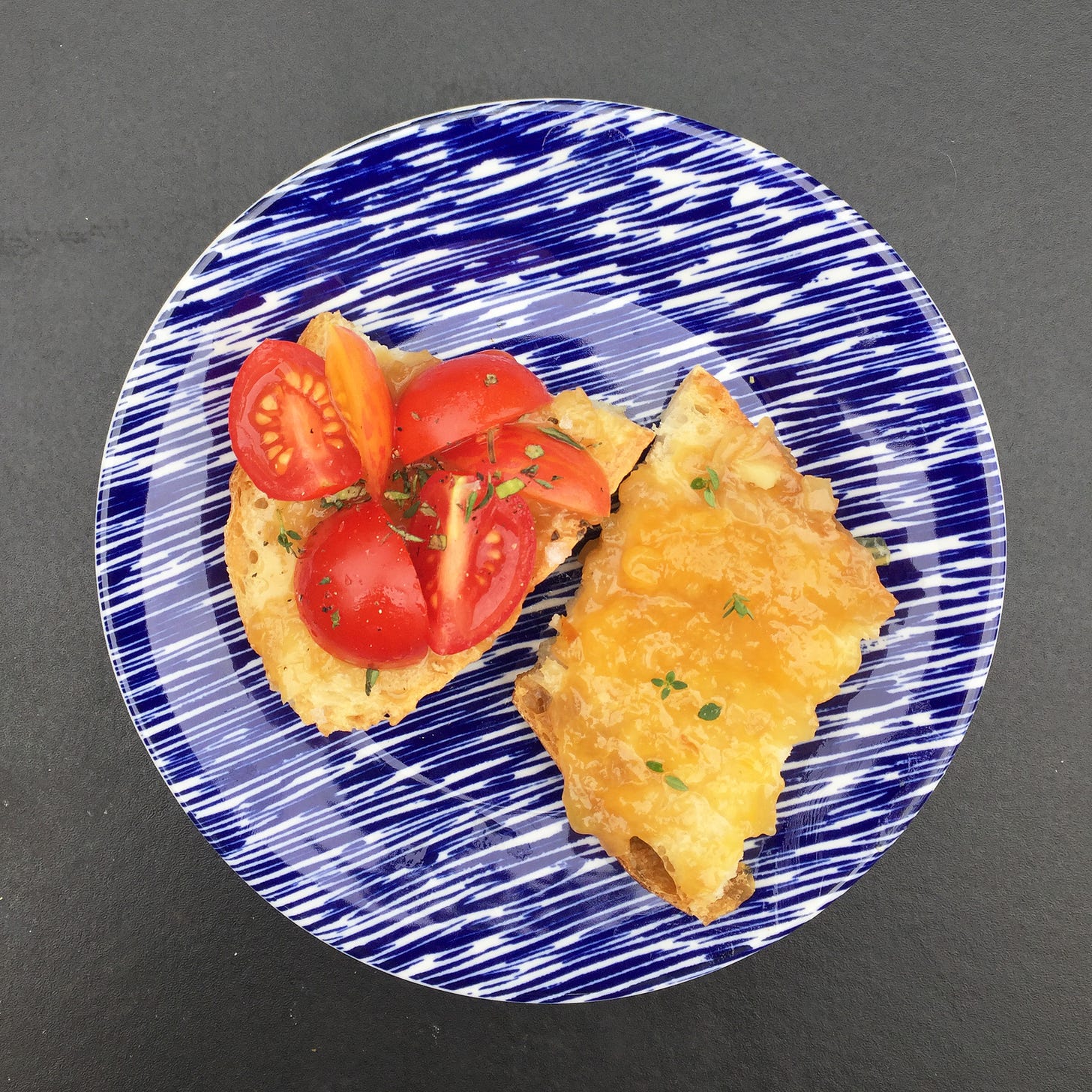 On a small blue plate, two half-slices of bread. On one is peach jam with a few thyme leaves, and on the other are quartered cherry tomatoes and chopped oregano.