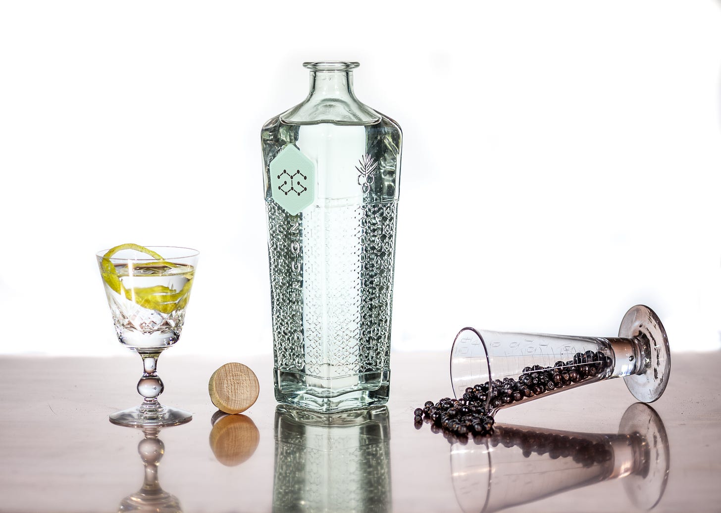 Image of a gin bottle on a tabletop, opened with its cork placed to its left. Further left is a martini in a goblet-shaped glass, garnished with a lemon twist. To the bottle's right is an upended glass of juniper berries, which are starting to spill out onto the tabletop. 