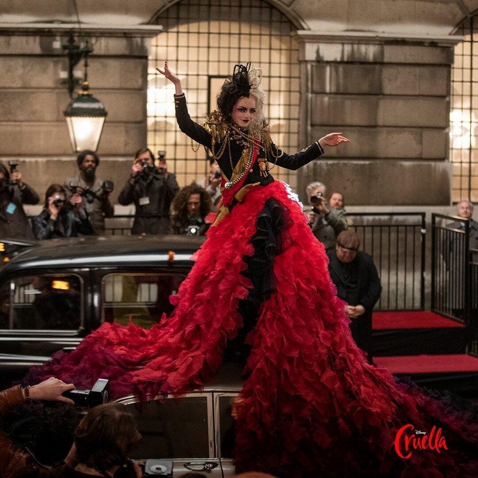 Cruella, played by Emma Stone, is posing with her arms out in a punk, embellished jacket, and flowing red skirt, atop a car in the street, while onlookers photograph her.
