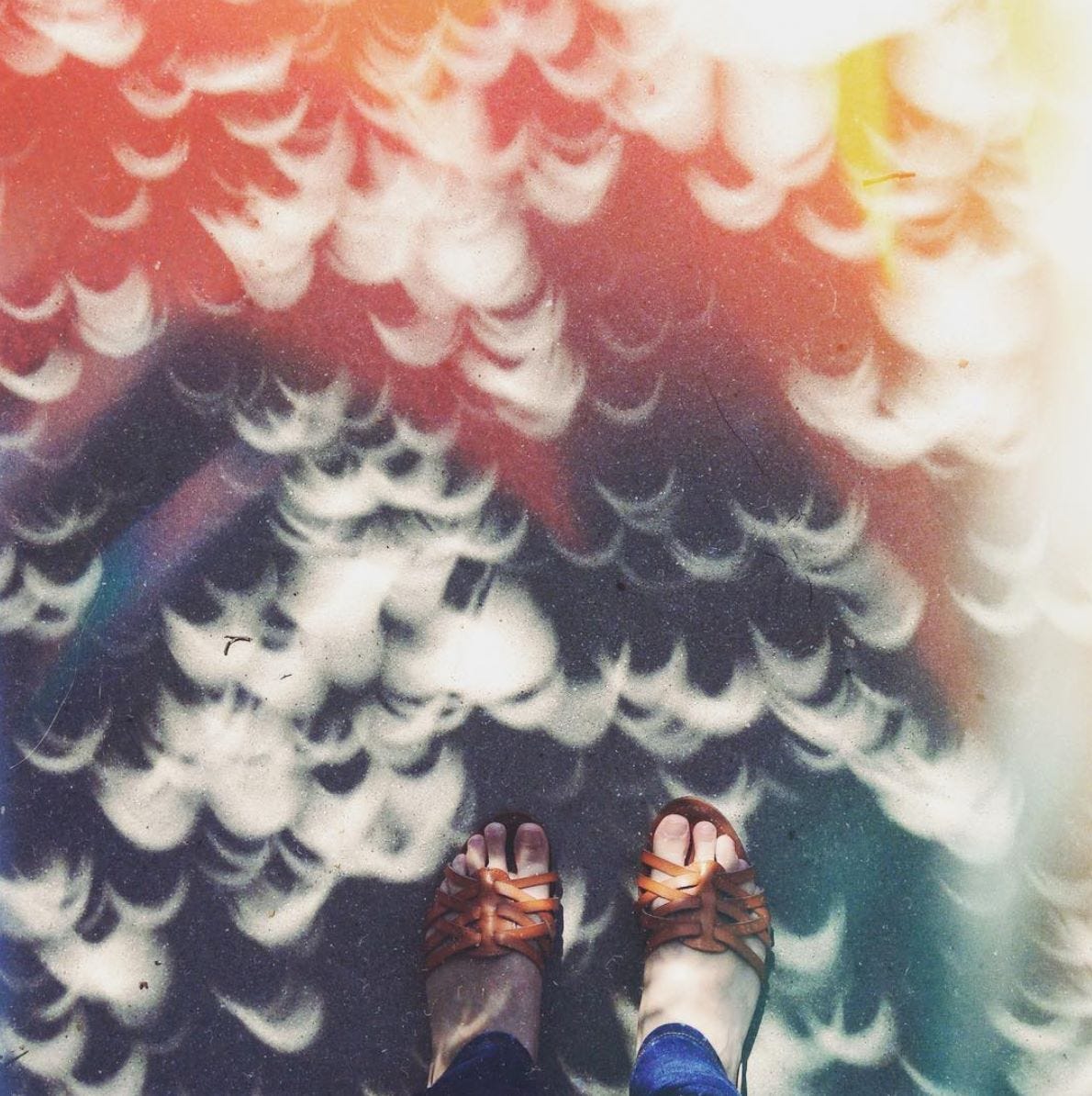 Feet in a pair of brown sandals and jeans are standing on a cement path with hundreds of suns from a solar eclipse. The filter is pink and yellow and orange.