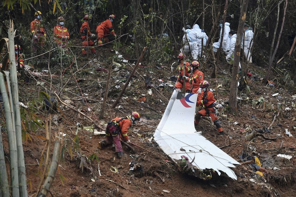 Several people wearing bright orange jackets and red helmets examine a piece of an airplane in a wooded area.