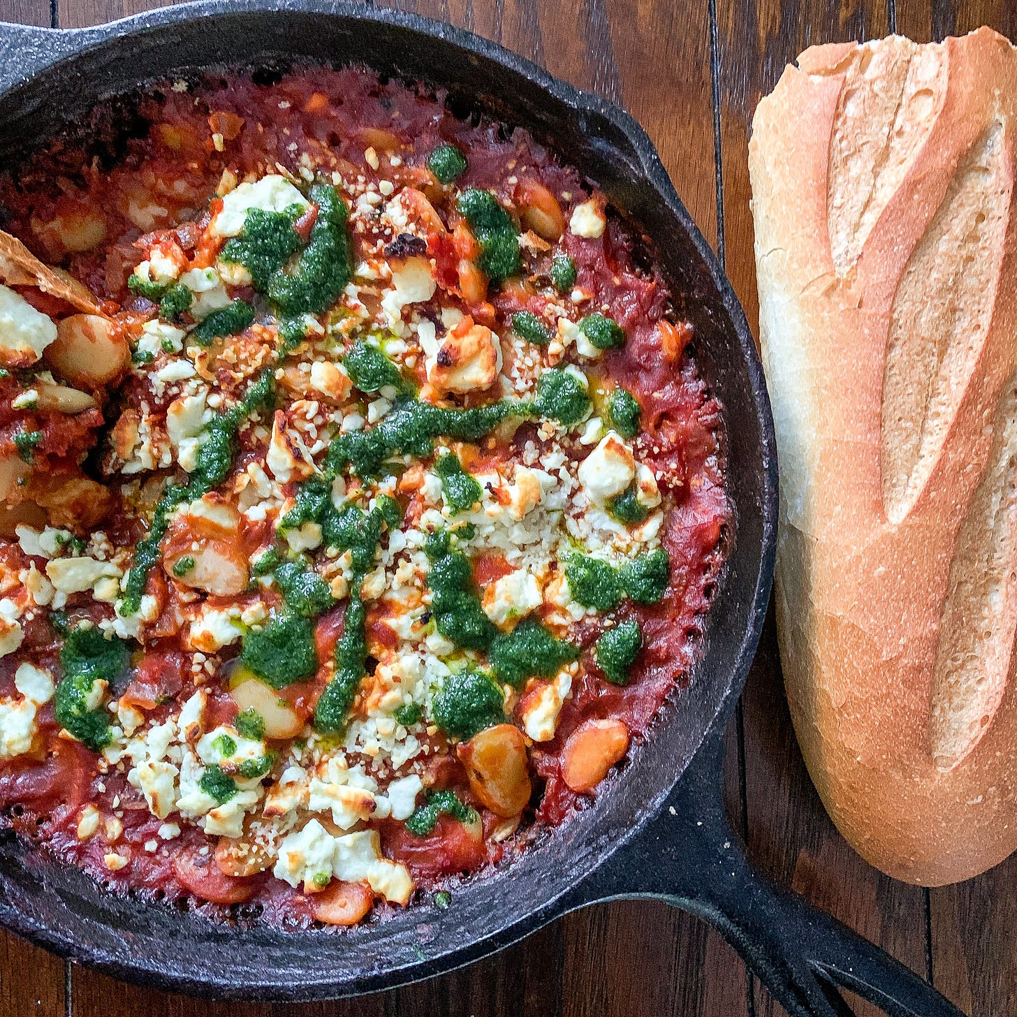 Cast iron pan on the left side is filled with a deep red and caramelized tomato sauce, spotted with white lima beans, topped with browned crumbles of feta, and drizzled with a bright green herb sauce. Half a rustic baguette is served to the right of the pan.