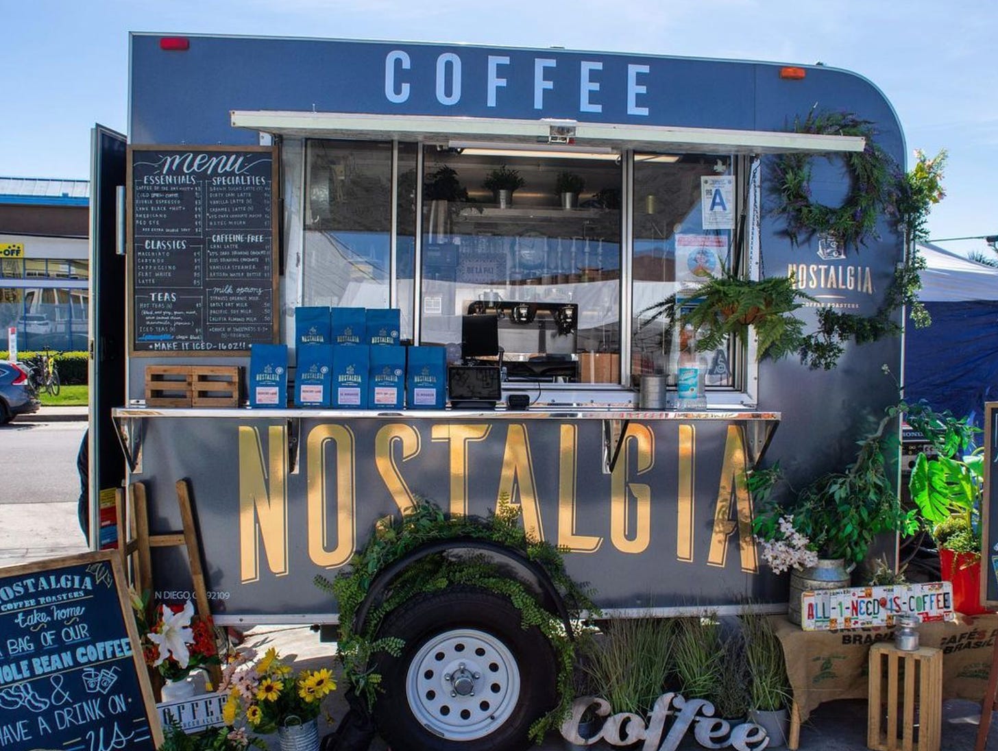 A coffee shop built into a pull-behind trailer. Photo is of the shop set up on the side of the road with blue coffee bags on the drop down counter, a chalboard menu mounted on the door and a slide open, walk-up window looking into the shops coffee bar with espresso machine.