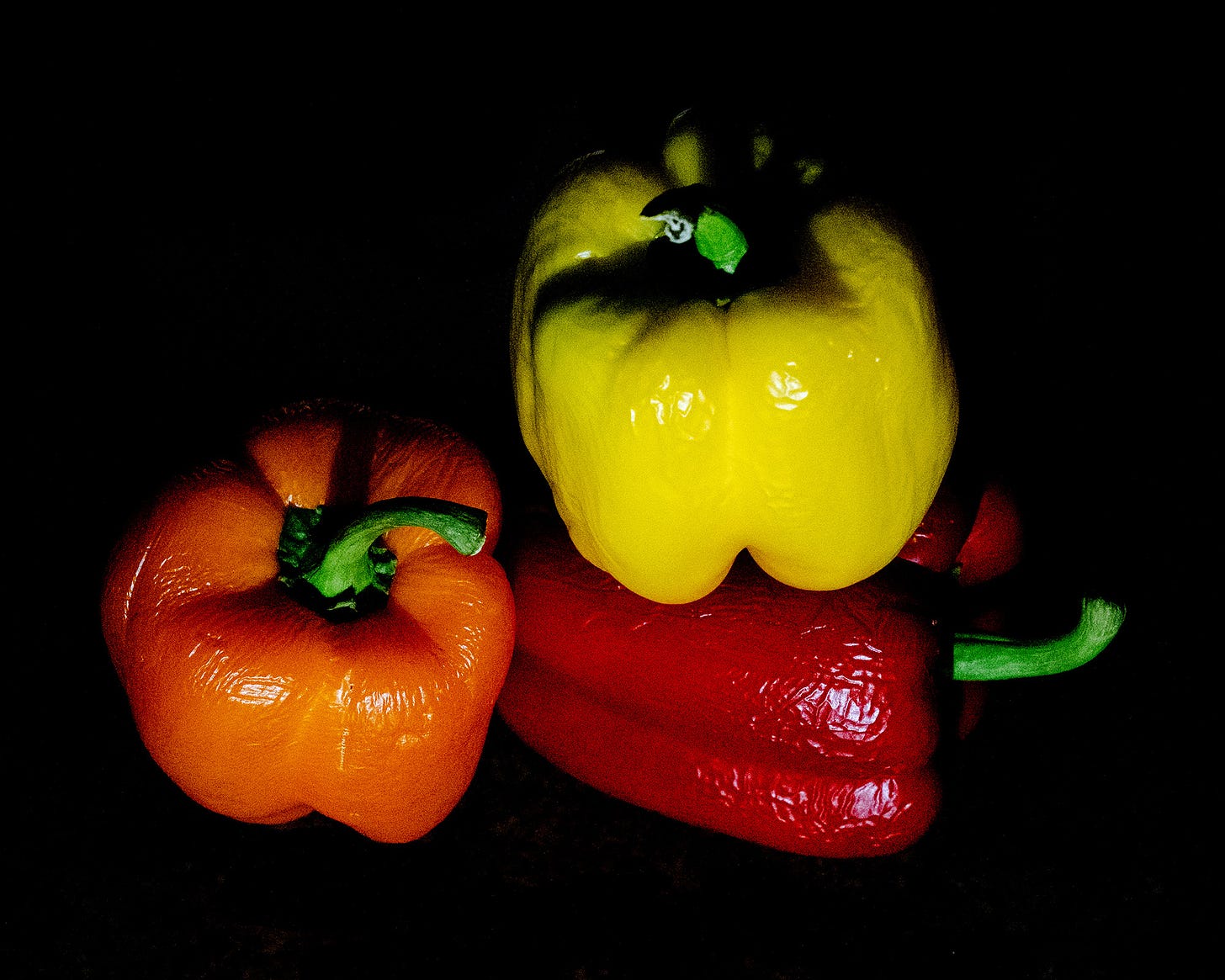 Three peppers - 1 red, 1 orange, and 1 yellow - arranged on a kitchen counter engulfed in shadows.