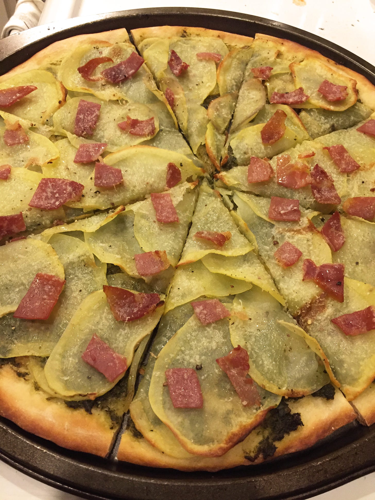 a pizza covered in thin slices of potato and diced prosciutto. Beneath the potatoes, a base of pesto is just visible.