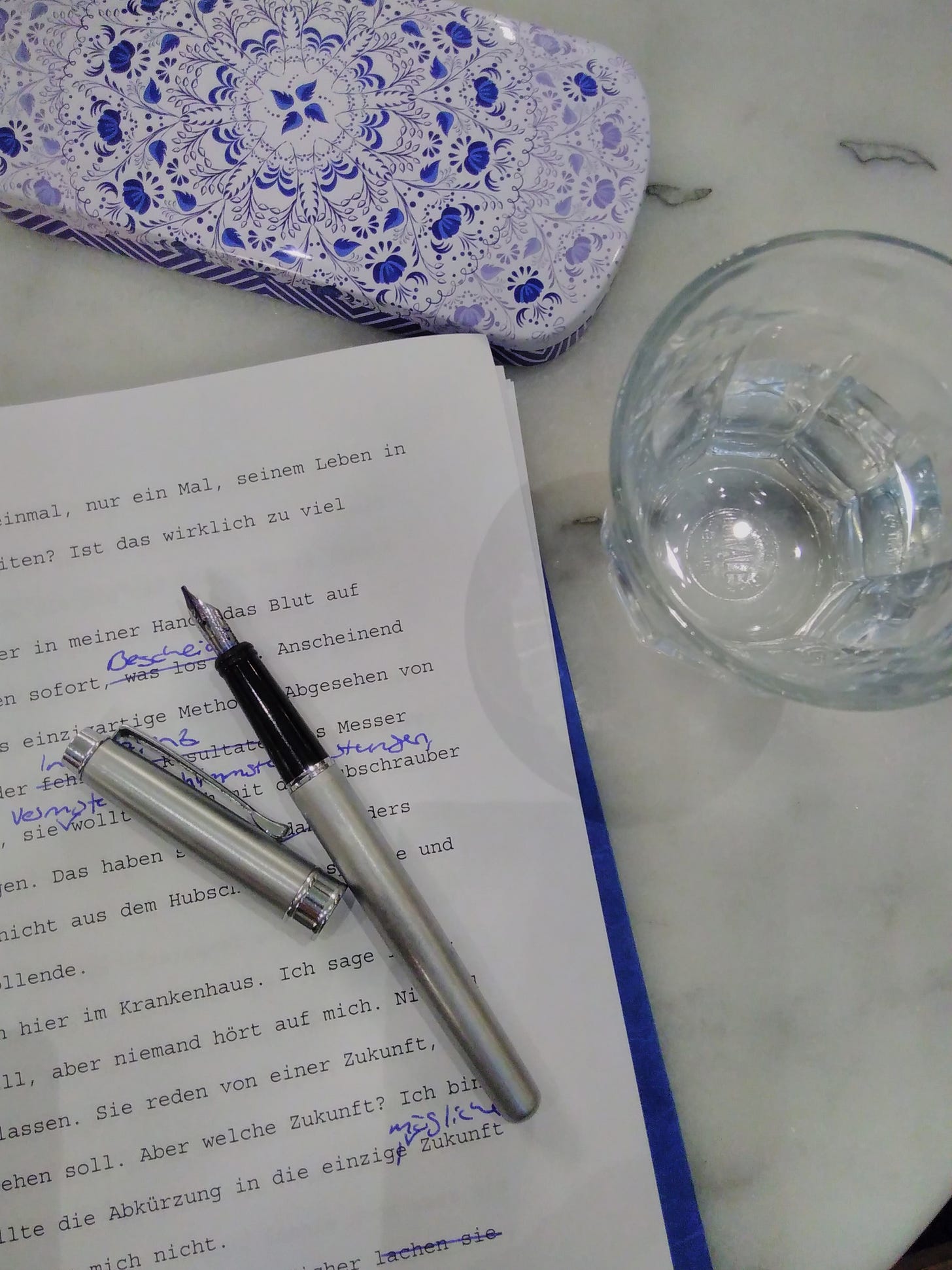 Ausgedruckter Text mit Korrekturen, darauf liegt ein silberner Füller, daneben ein leeres Glas und ein Federmäppchen aus Blech mit blau-weißem Muster. Die Tischplatte ist aus Marmor.