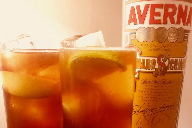 Two glasses full of amber liquid, ice cubes, and lemon slices next to a bottle of Averna amaro.