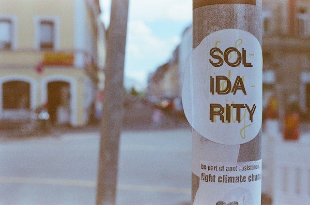 In the foreground, a sticker on a post with "Solidarity" in black capital letters superimposed over the same word written in lower-case cursive yellow letters. Blurred in the background is a street with a yellow building.