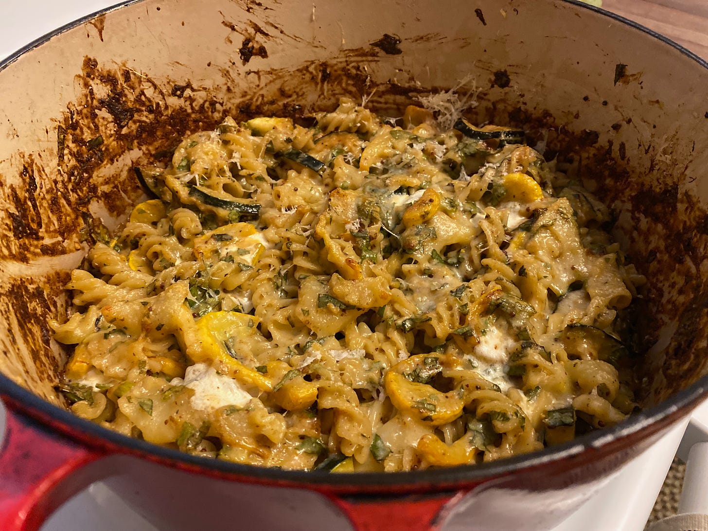 A large Dutch oven holding a cheesy pasta bake with pools of melted mozzarella, specks of fresh herbs, and crisped brown summer squash and zucchini slices.