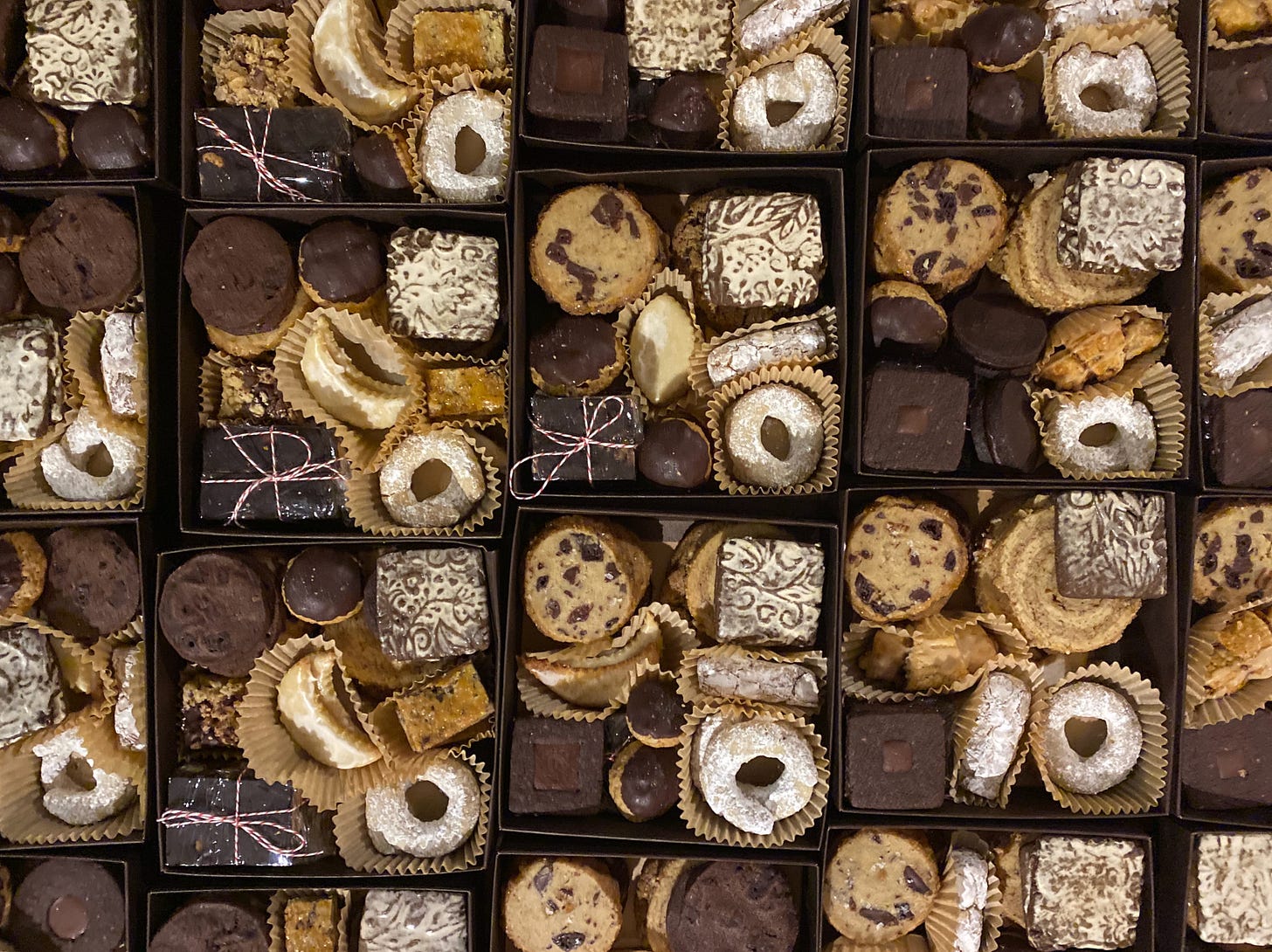 A closeup of many small square cookie boxes in tight rows. Each box contains the same cookies, a mix of round shortbreads, crescent shaped cookies, rings coated in powdered sugar, square chocolate cookies, rugelach, and others.