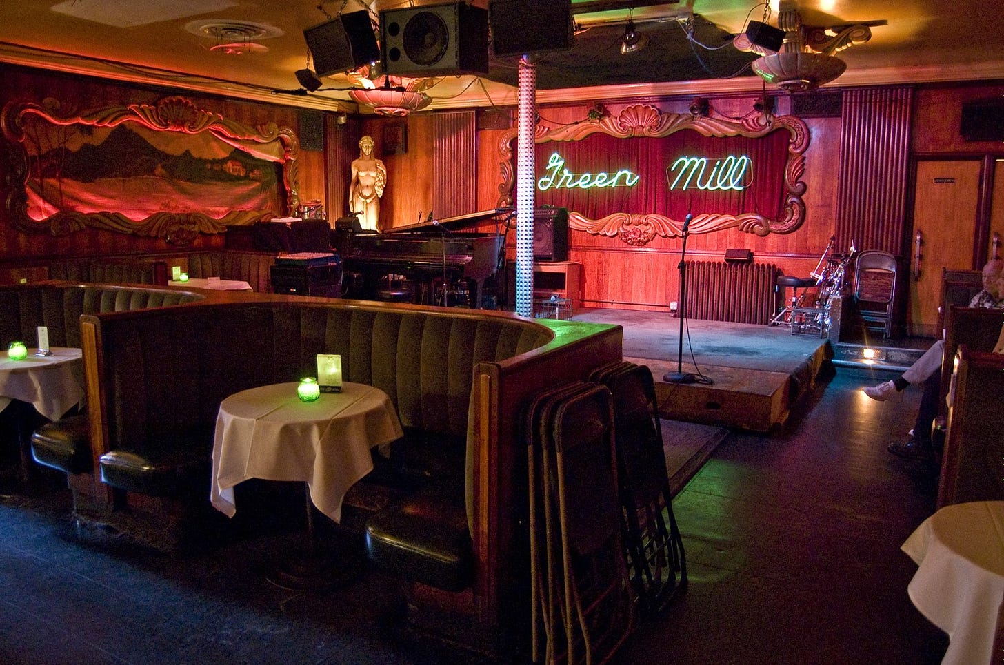 A view inside a nearly empty Prohibition-era night club, with U-shaped padded booths facing away from a small stage.