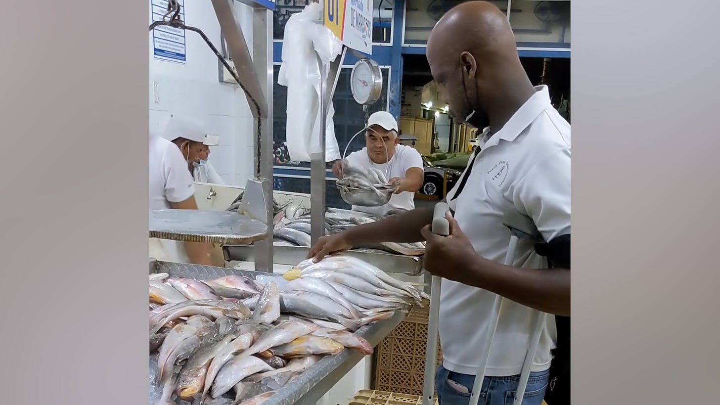 A man wearing a white polo shirt and blue jeans is touching a fish on a table full of whole fish for sale.