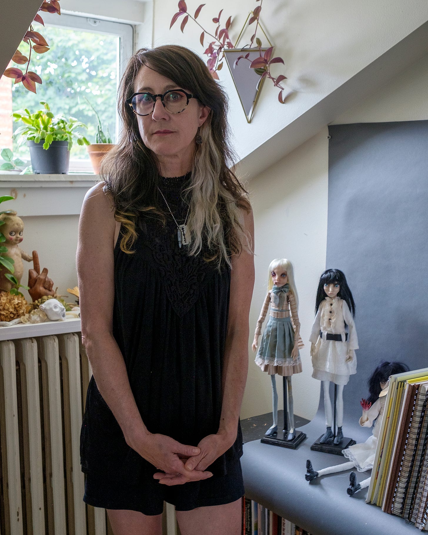 Beth Robinson stands in front of a window with two strange dolls at her side. One doll has blond hair and the other black hair.