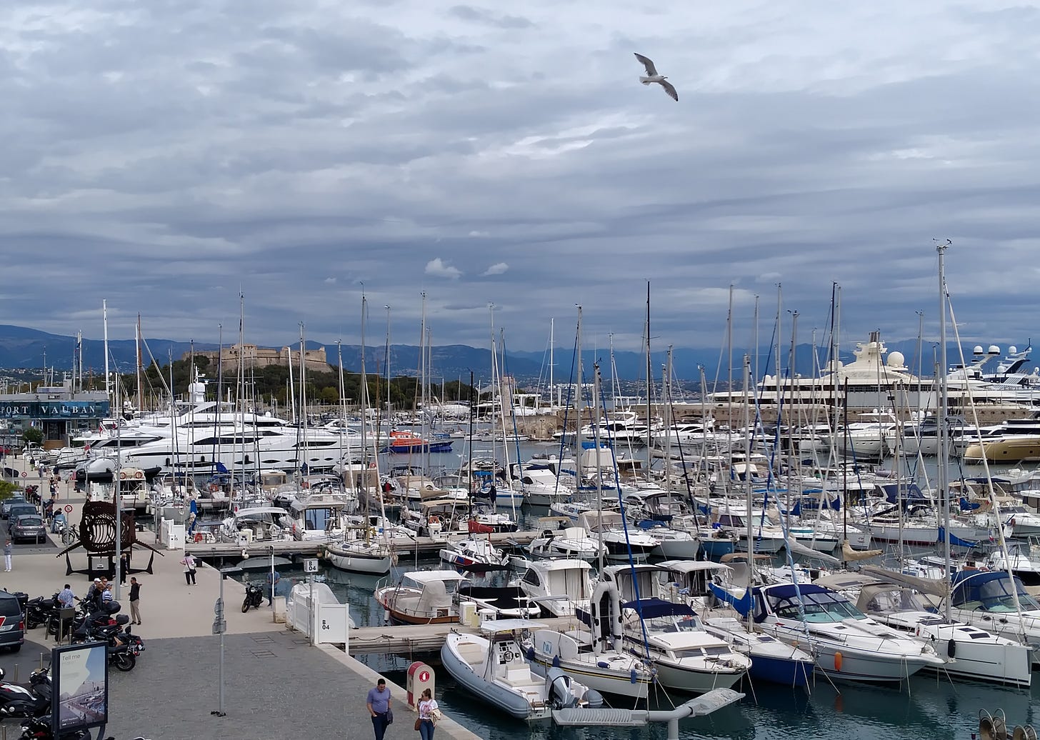 photo of yachts in the harbor at Antibes, France. The day is bright but still cloudy, the moorings are full. In the background some mega yachts and in the far distance an old fort on a hill. 