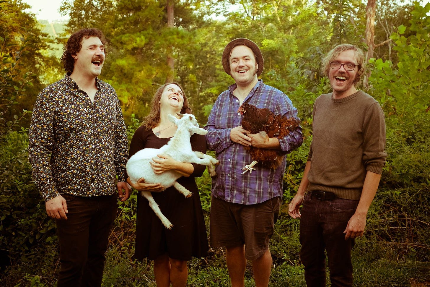 Four people stand in a forest setting. From left to right, there is a white man wearing a tightly patterned floral shirt. His eyes are nearly closed and he is laughing, and he has curly brown hair and a mustache. Next is a young woman, laughing, holding her head back, holding a white goat. She is wearing a burgundy dress and has light brown hair. Next is a man with a blue flannel shirt, wearing a brown hat, holding a brown chicken. He is smiling, eyes off the camera. Second is a white man wearing a brown sweater with the sleeves rolled up. He has blonde hair and is wearing glasses, looking directly at the camera. This is their label promo photo.