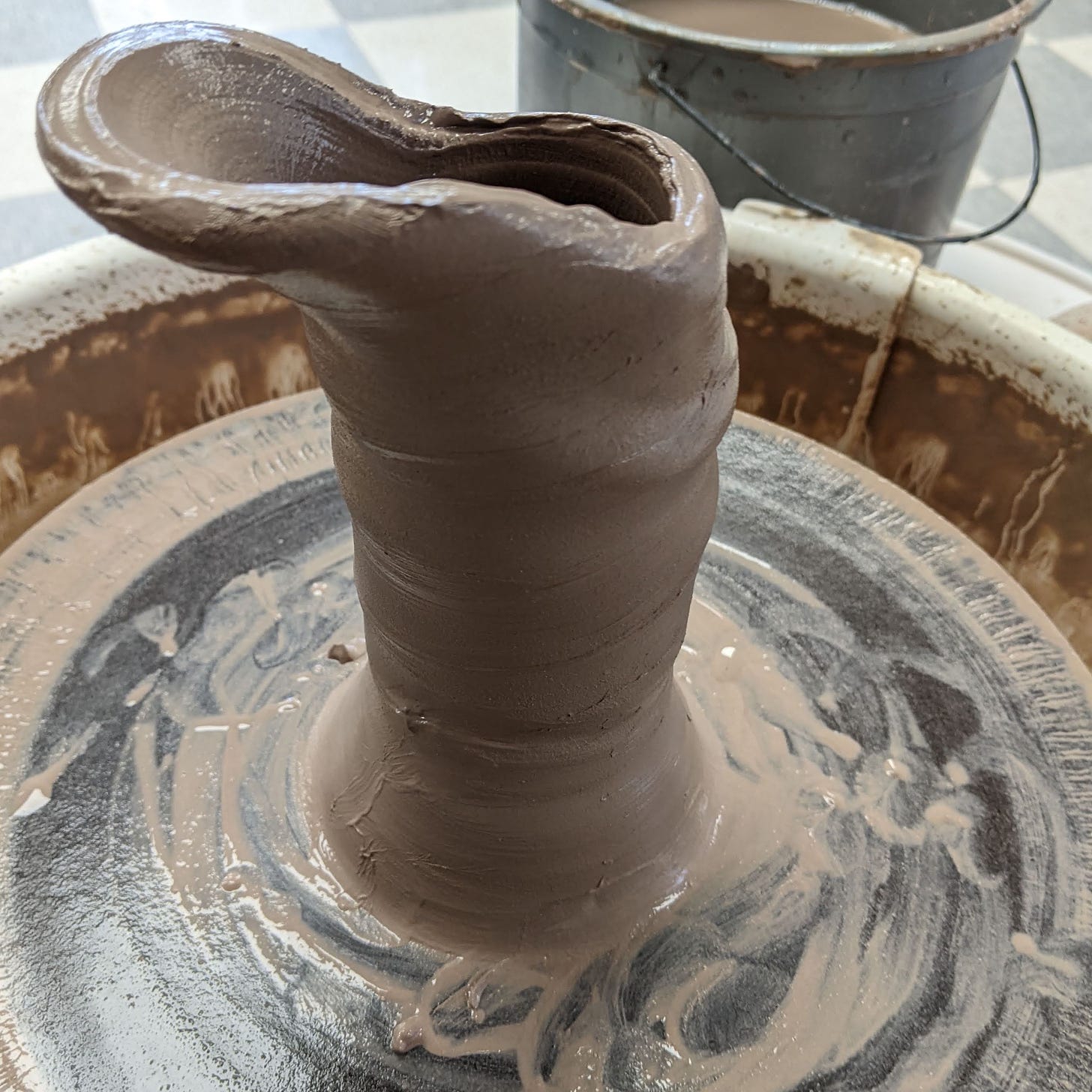 Wonky failed attempt at a pottery cylinder, with a spouty looking edge off to the side, on a wet bat, on a pottery wheel