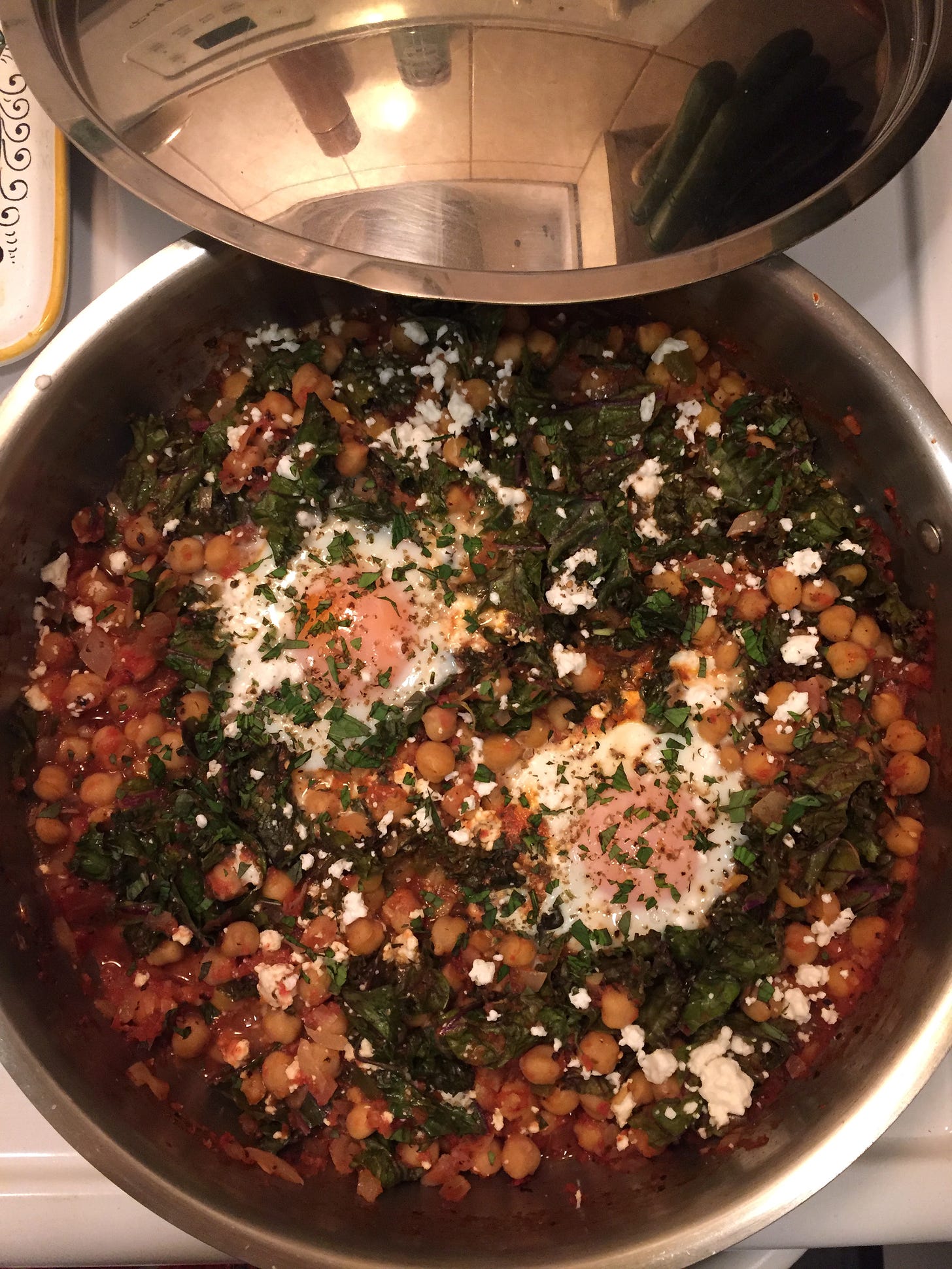 A large stainless steel pan full of the chickpea and kale shakshuka, with two poached eggs in the centre. The top is scattered with feta, mint, oregano, and za'atar.