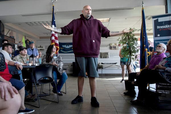 John Fetterman speaking at an event in May, wearing a burgundy hooded sweatshirt, gray athletic shorts and black sneakers.