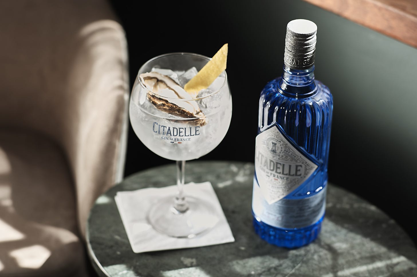 A small bottle of Citadelle gin on a table next to a glace with ice, a twist of lemon, and a single oyster. The color palette is muted grays and pewters, but the gin bottle is a light clear ceylon blue.