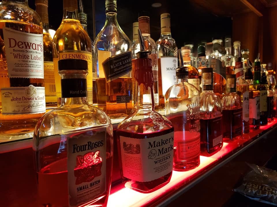 A whiskey bar with rows of whiskey bottles in tiers lighted from beneath. There's a Four Roses, Maker's Mark, Dewar's White Lebal, Dewar's black label and more.