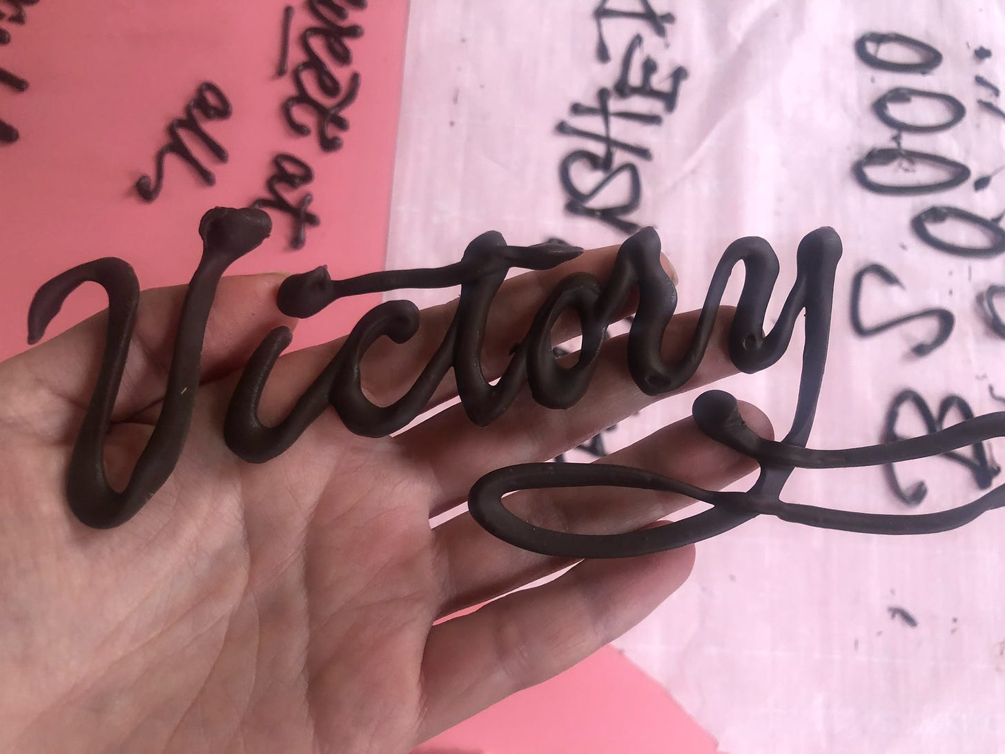 A hand holds "victory" lettered in untempered, dull chocolate over a parchment paper work surface with additional chocolate letters.