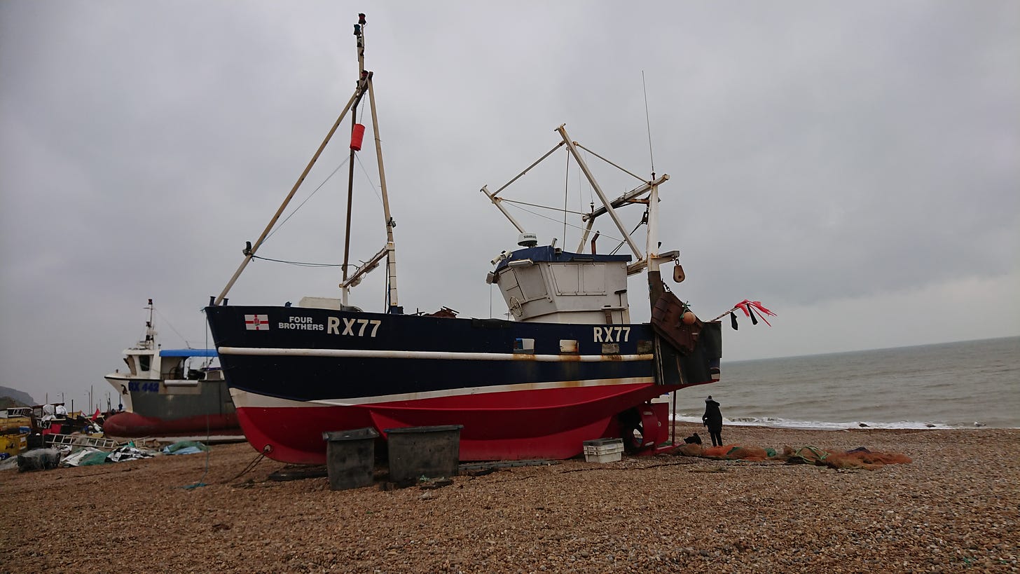 A boat with a red bottom, white stripe, and blue top sits against the backdrop of an overcast grey sky. The sea comes in at an angle just behind it, with the stony ground in the foreground.