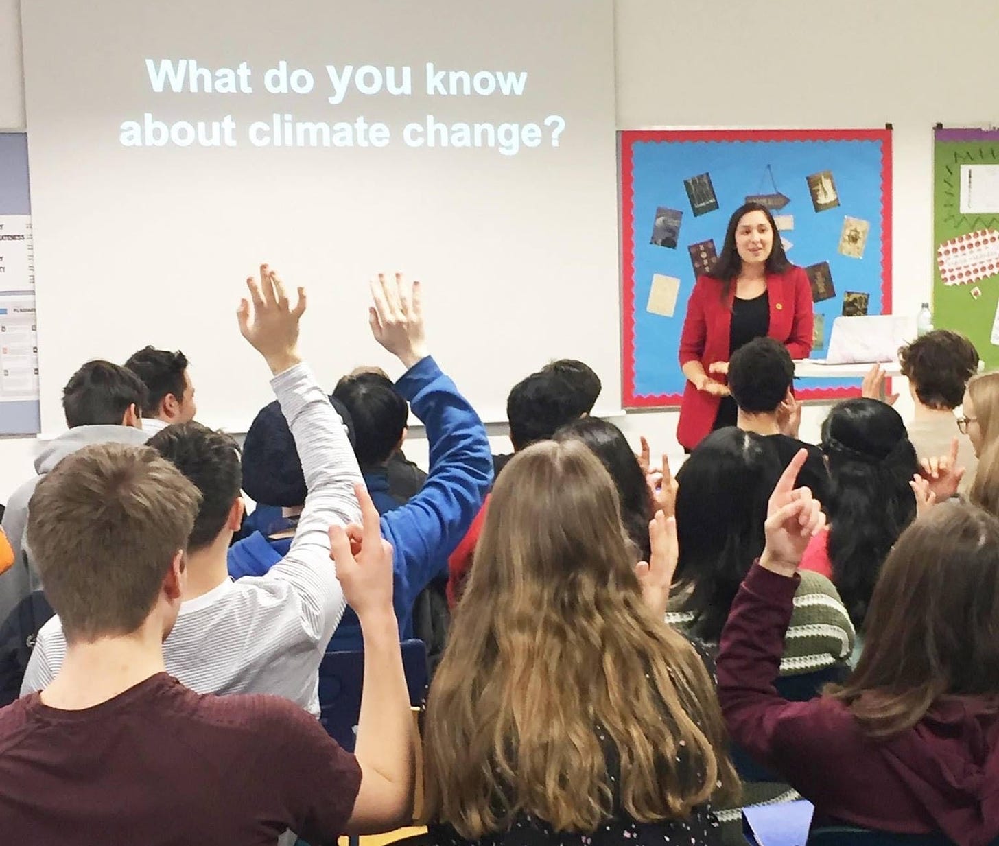 Bradlie stands at the front of a classroom of young teens by a Powerpoint slide that says 'What do YOU know about climate change?'