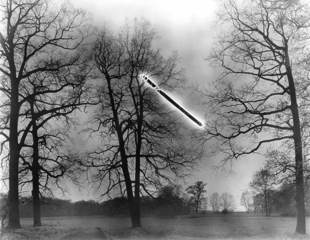 fotografia em preto e branco de um campo com árvores e um bastão negro brilhante flutuando no ar entre as árvores