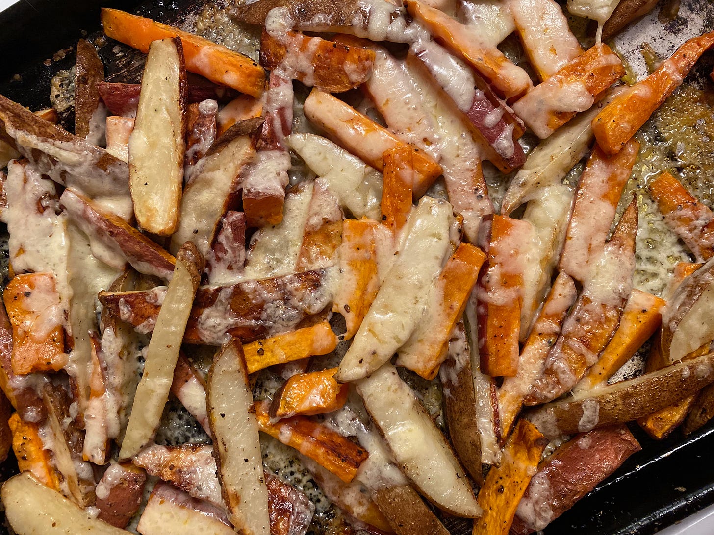 A pile of white and sweet potato wedges on a baking tray, covered in cheese. The potatoes ave crispy brown edges, and the cheese is melted and bubbling.