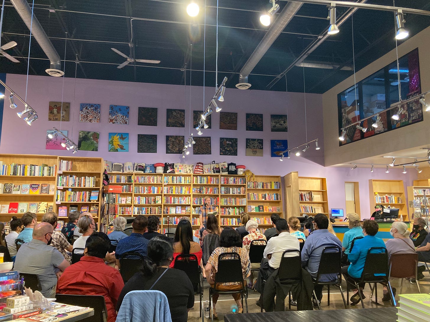 A brightly colored bookstore hosts a sizable crowd of listeners as Chris Martin reads from a book. 