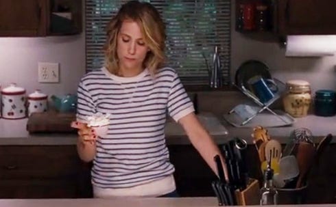 A medium shot of a woman standing in a kitchen. She's wearing a blue and white striped shirt and is holding a cupcake in one hand, looking down at it.