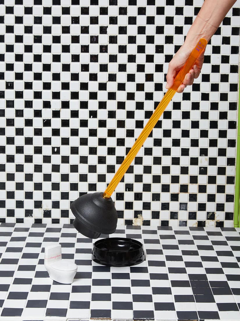 A black and white tiled bathroom with someone holding a plunger that has an orange acrylic handle 